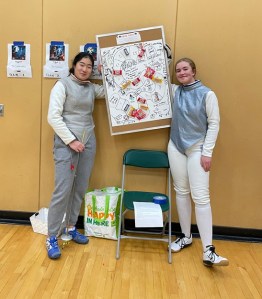 Two foil fencers stand with the halloween challenge prize wall
